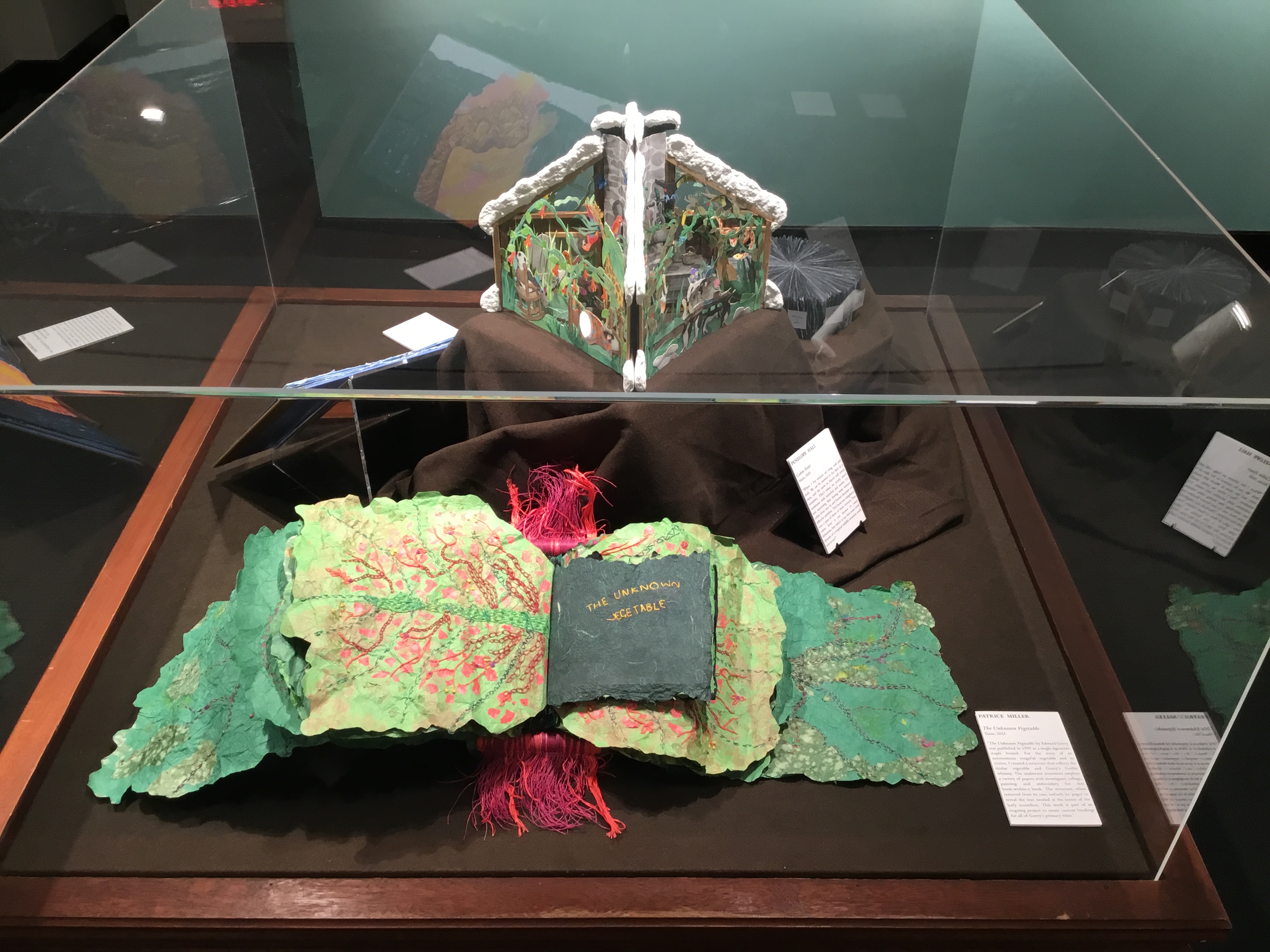 A plexiglass floor case in the small gallery displaying several books: the foremost book, a salad of leafy green paper pages flopping out beside an interior page inscribed with “The Unknown Vegetable” in gold, is bound together by a mass of red threads.  Behind it on a pedestal stands a pop-up carousel book depicting colorful tropical animals bound together in a paper cabin topped with snow.