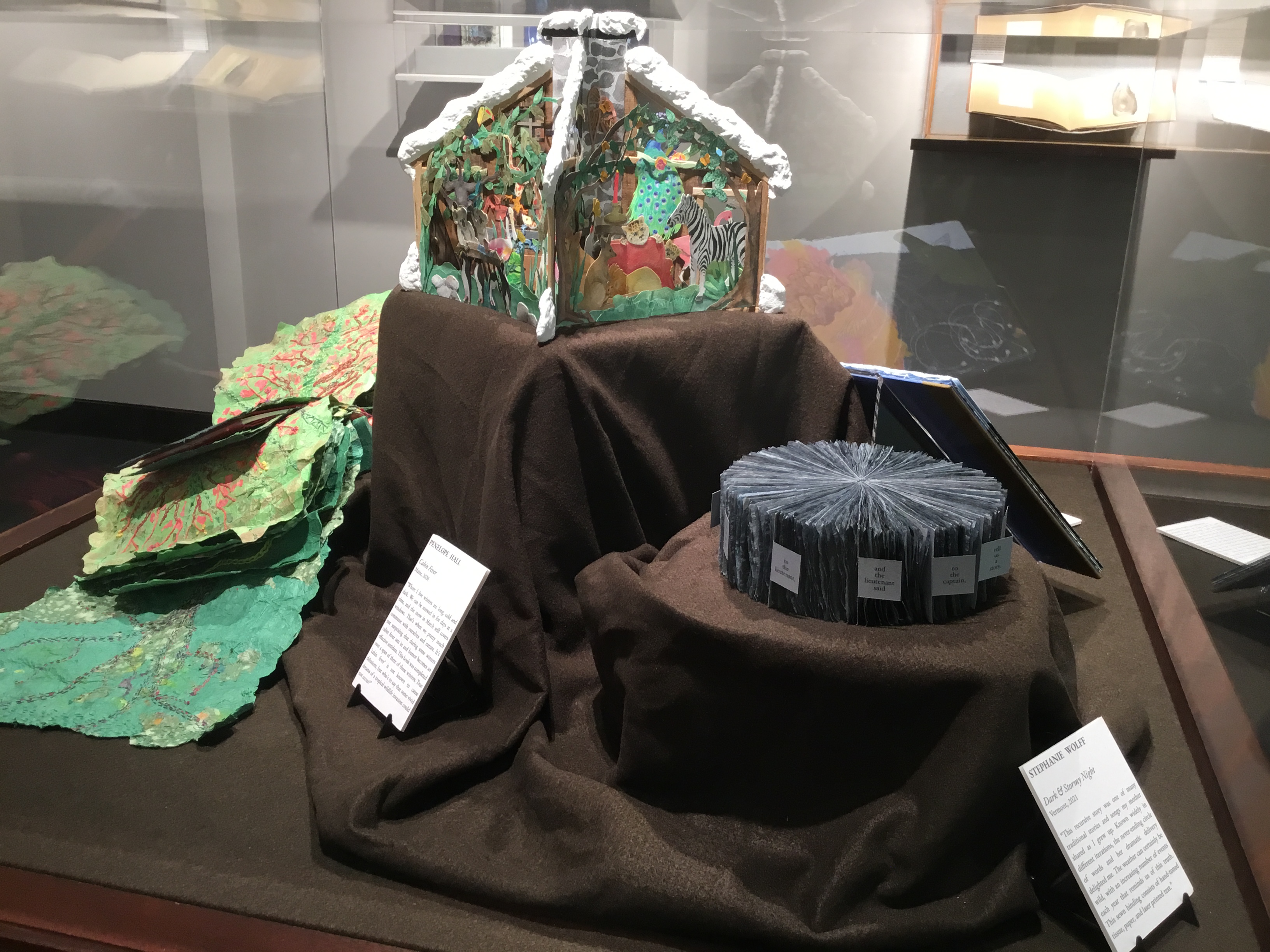 A plexiglass floor case in the small gallery displaying several books. On the left is a salad-like book of leafy green paper pages embroidered with veins and flopping out beside an interior pages bound together by a mass of red threads.  Behind it on a pedestal in the middle stands a pop-up carousel book depicting colorful tropical animals bound together in a paper cabin topped with snow.  The right foreground pedestal displays a blueish gray book that forms a cylindrical shape bound at the center.