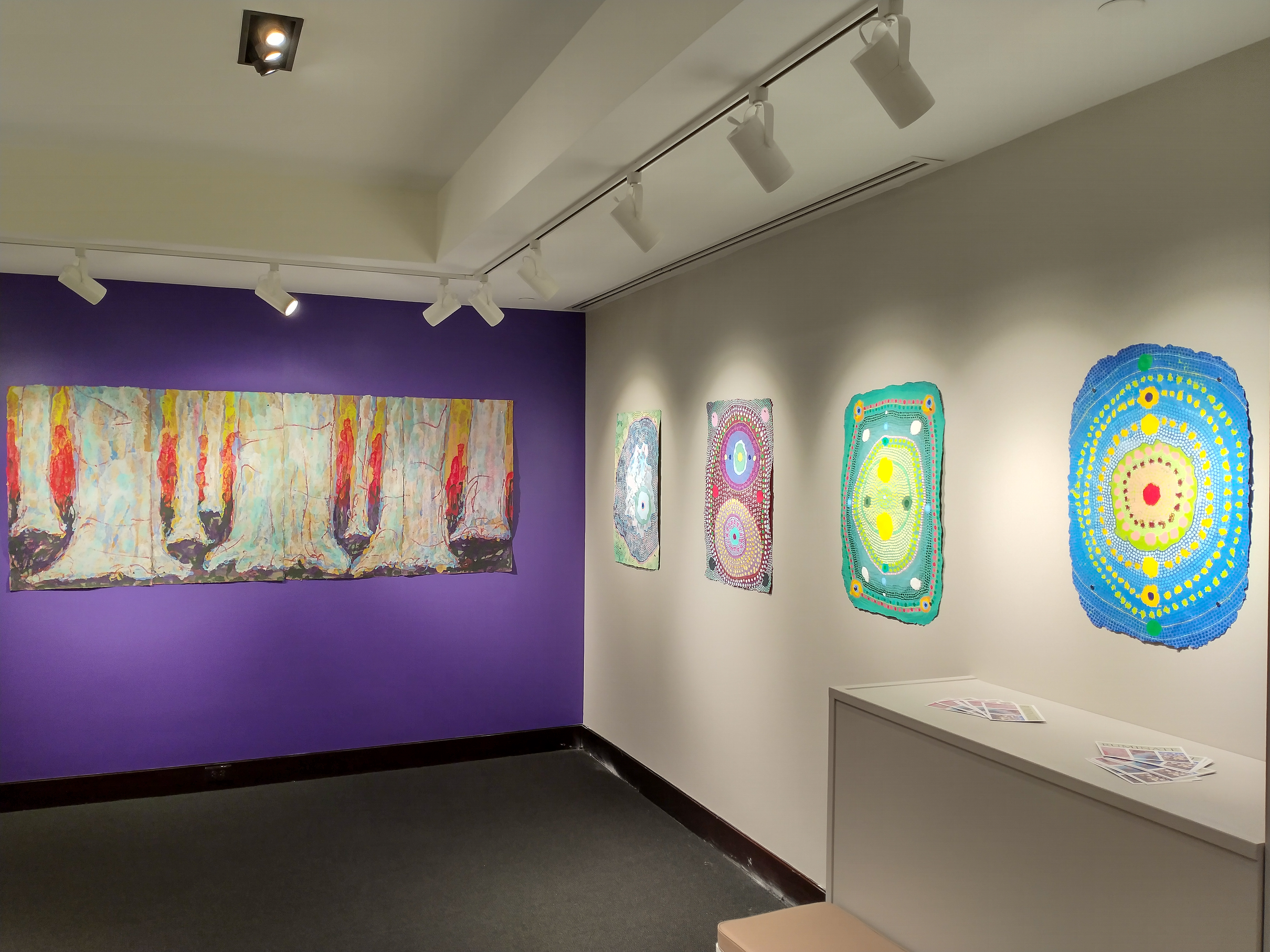 View of the east wall of the small gallery. All the pieces on this wall are created by Chad Hayward. From right to left, the order in which you would encounter the pieces upon entering the gallery: Gateway is a primarily blue piece with concentric circles radiating from a red dot at the center of the page. The concentric circles are created by a line of smaller circles. Papillon is a rectangular green piece with two yellow dots stacked one above the other at the center of the piece and two dark green dots arranged horizontally. These two sets of dots organized in cardinal directions sits atop a field of small white and yellow dots arranged radially in concentric circles. A rectangle of red dots runs along the outer edge of the piece. Element 115 is another series of radial dots on a rectangular sheet the colors lean more towards purple, yellow and white. The final piece, titled Rock Salt features the repetitive dot pattern like the other pieces but is shaped more organically. This piece features a blue form on top of a green background with the entire thing being coated in a light layer of white fibers.  