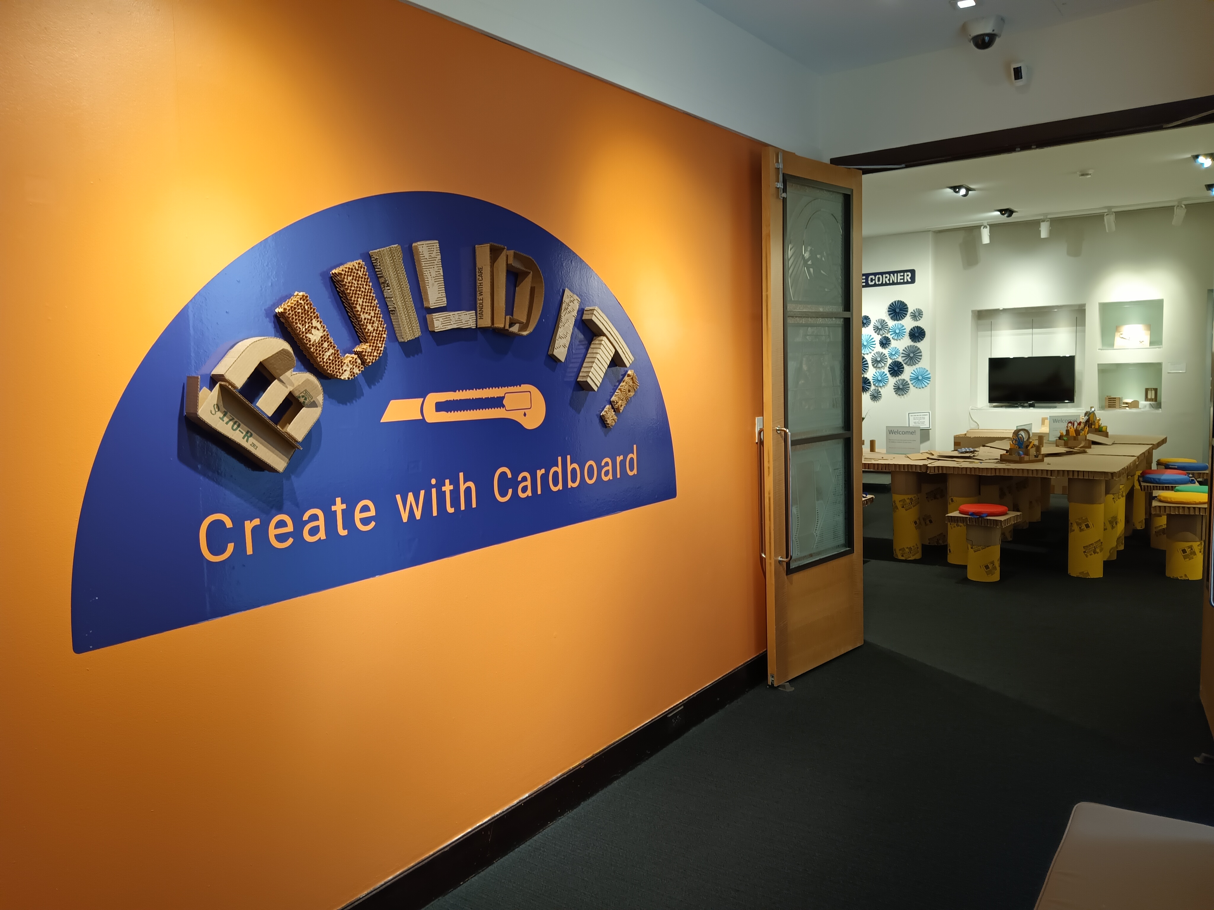 Photo of the entryway to Build It! Create with Cardboard. The title Build It, is sculpted from cardboard and placed in a deep purple semicircle on a bright orange background. The double doors to the right are open to the exhibit.