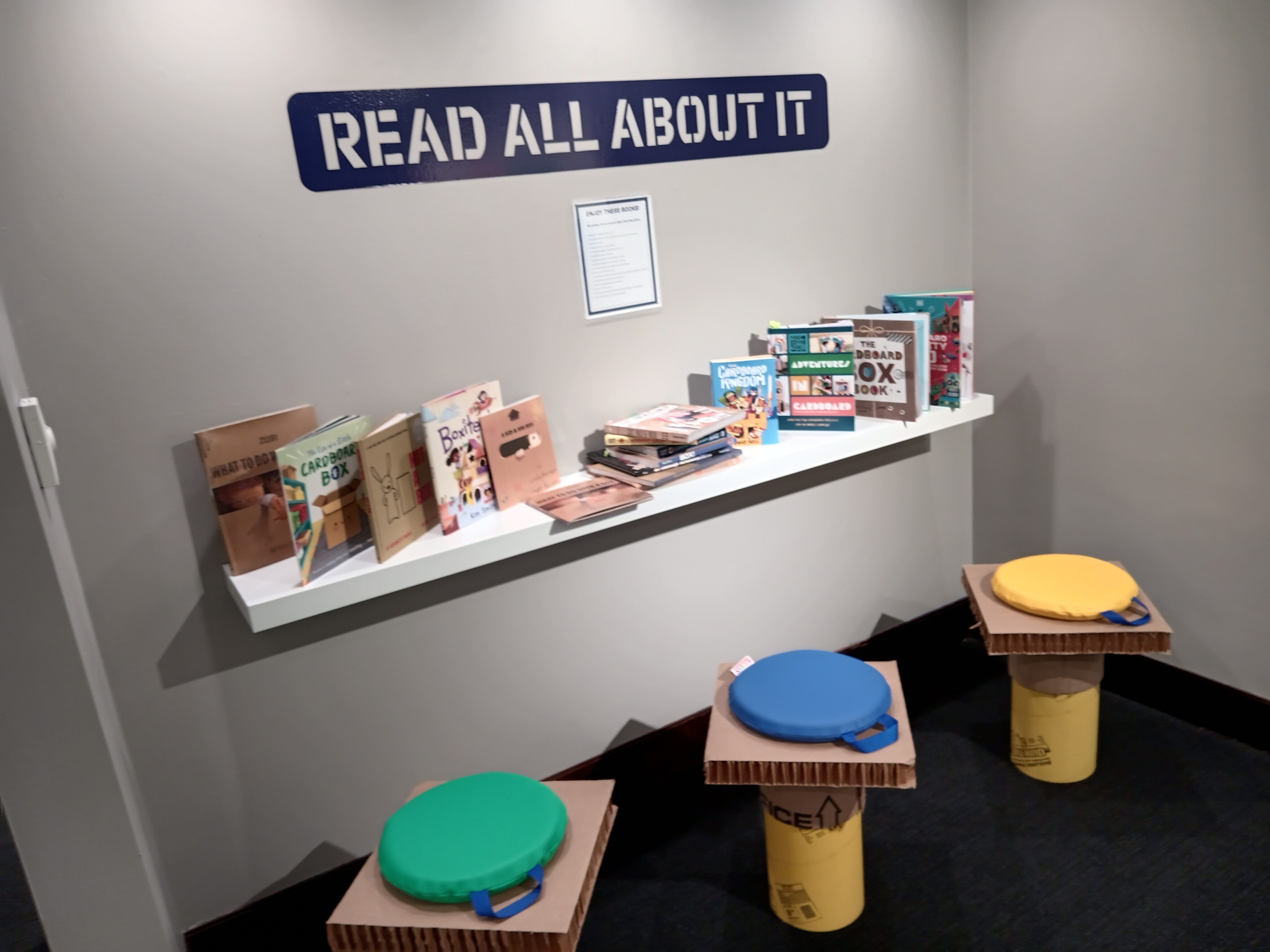 View of the reading corner in the back gallery. A sign announces Read All About It is displayed above a white shelf containing over a dozen books on paper and paper crafts. Three cardboard stools with colorful cushions are placed in front of the shelf.