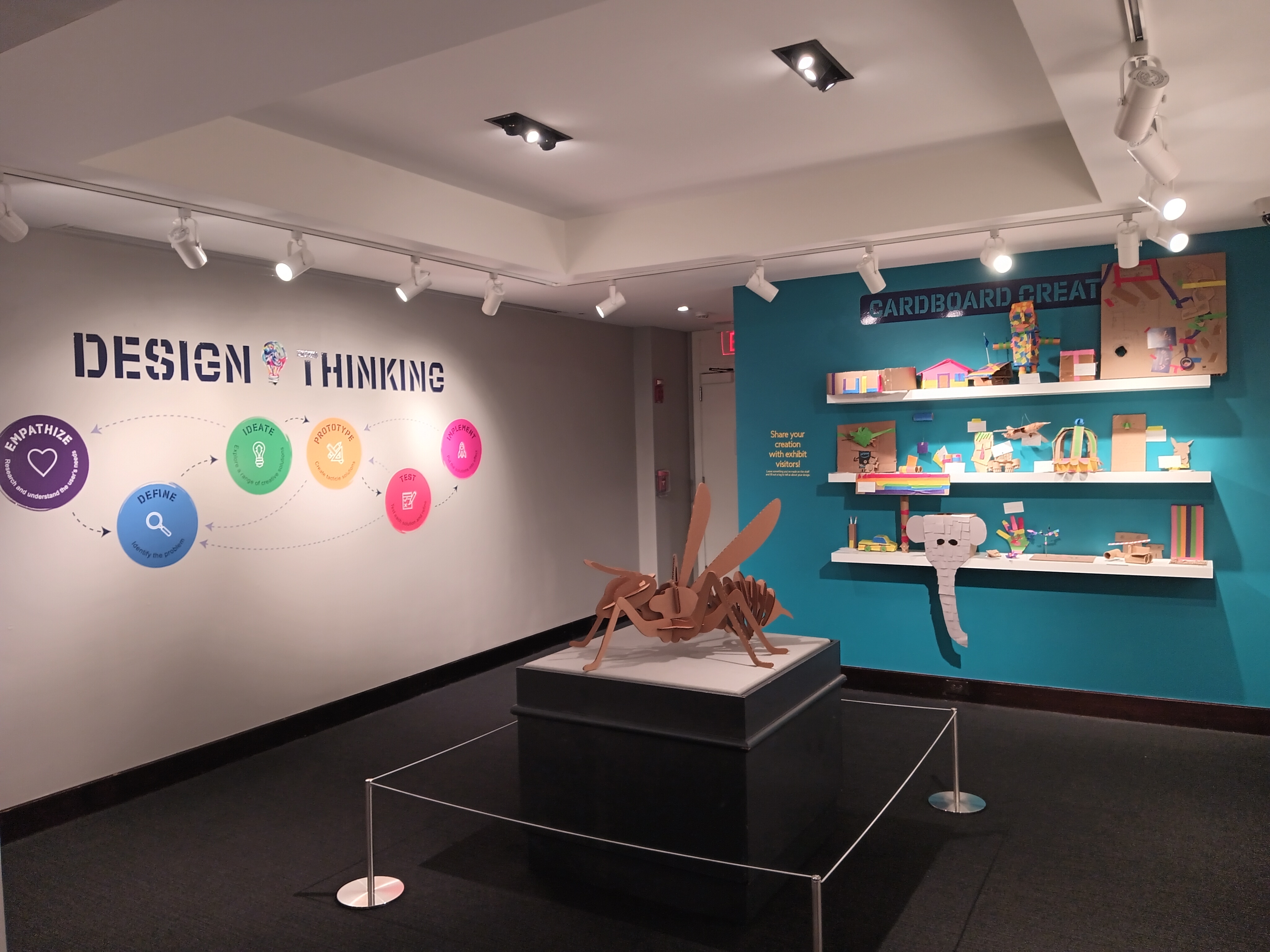 View of the back gallery. The left wall displays a large graphic titled Design Thinking, and colorful circles and arrows outlining the design process. In the center of the room, is a large cardboard yellow jacket on a black pedestal. The back wall is painted teal and labeled Cardboard Creations; and displays various cardboard sculptures crafted by museum guests on three white shelves.