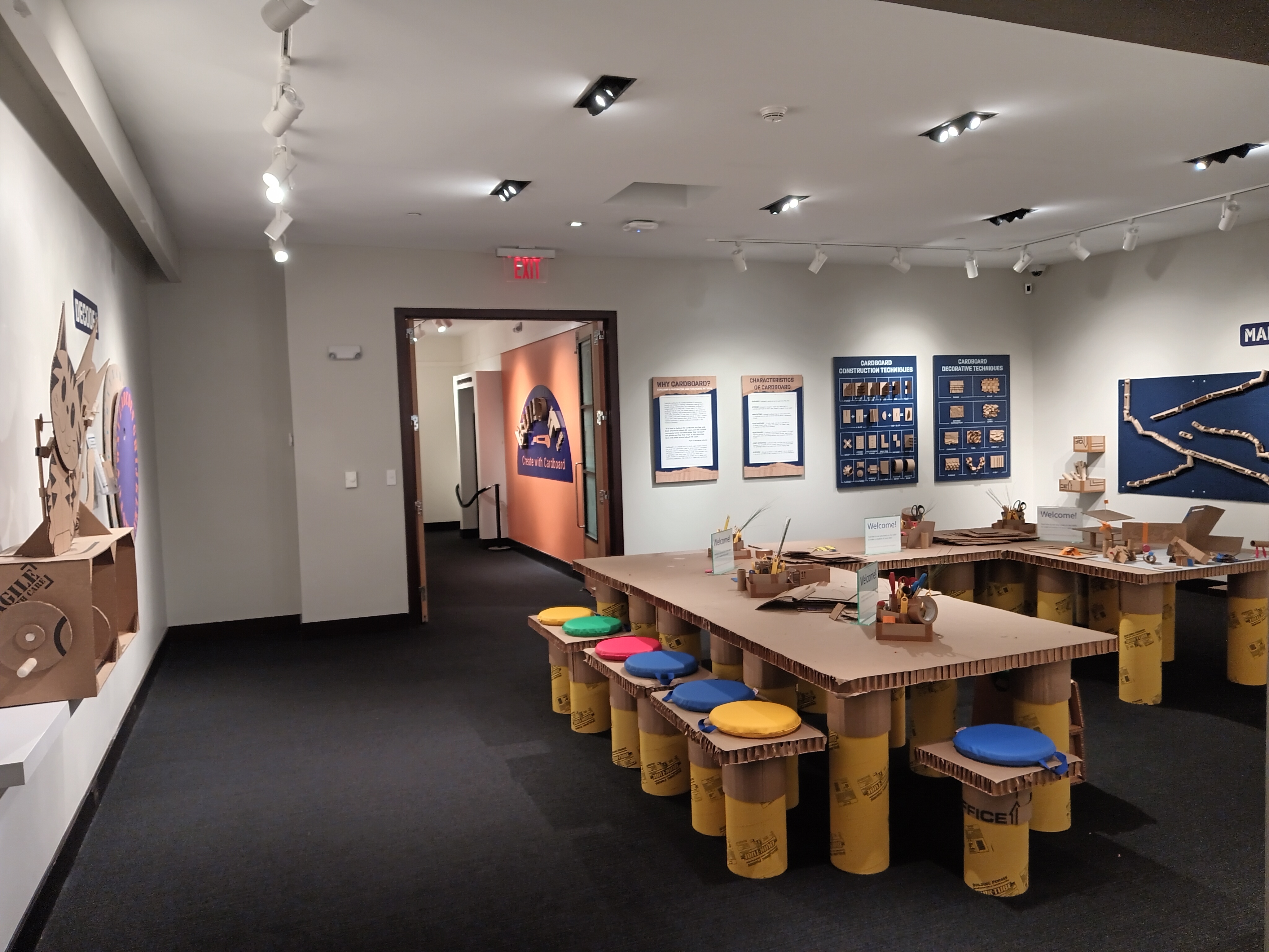 Overall view of the front gallery facing the entrance. In the foreground are cardboard tables, chairs, and various materials for guests to craft cardboard creations.