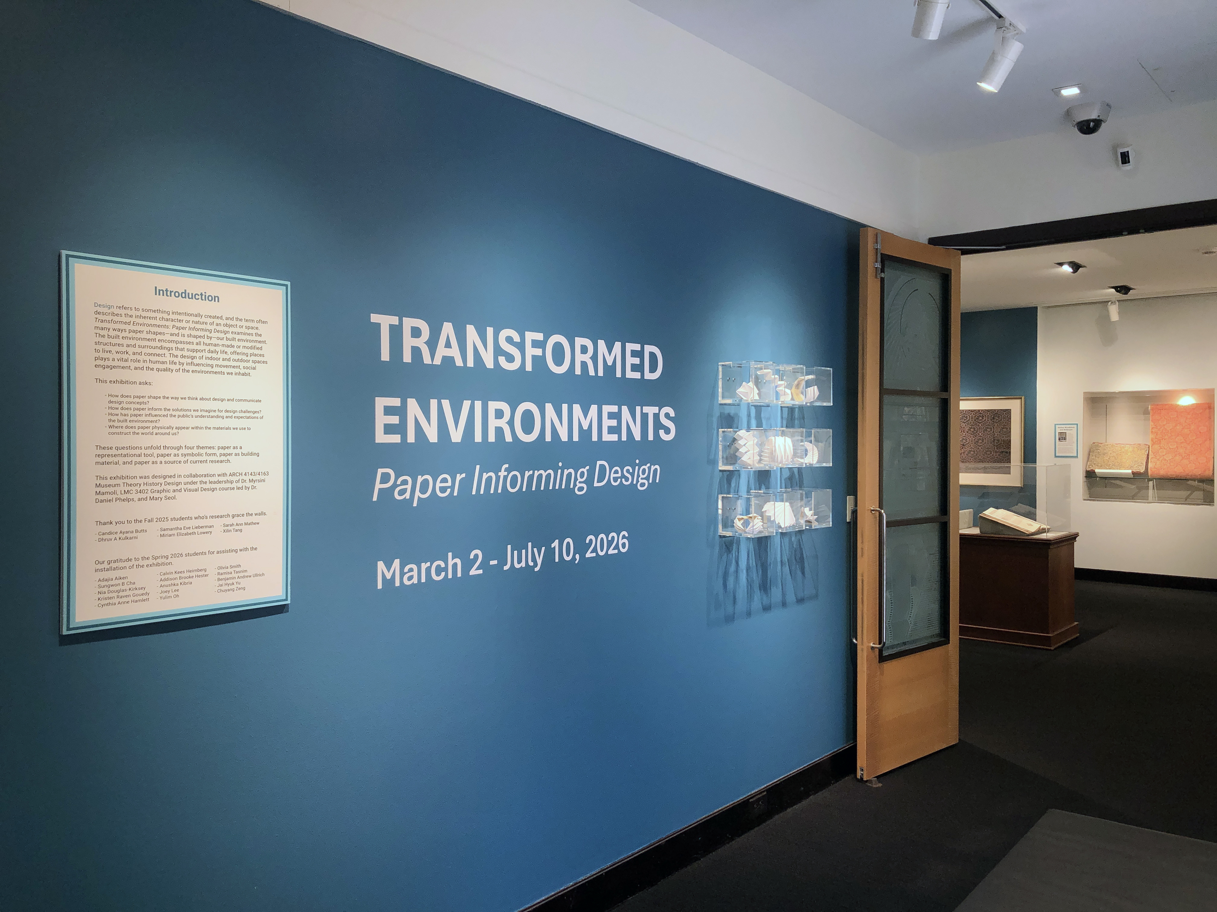 A blue wall with “Transformed Environments: Paper Informing Design” in white text with folded paper models mounted in plastic cases.