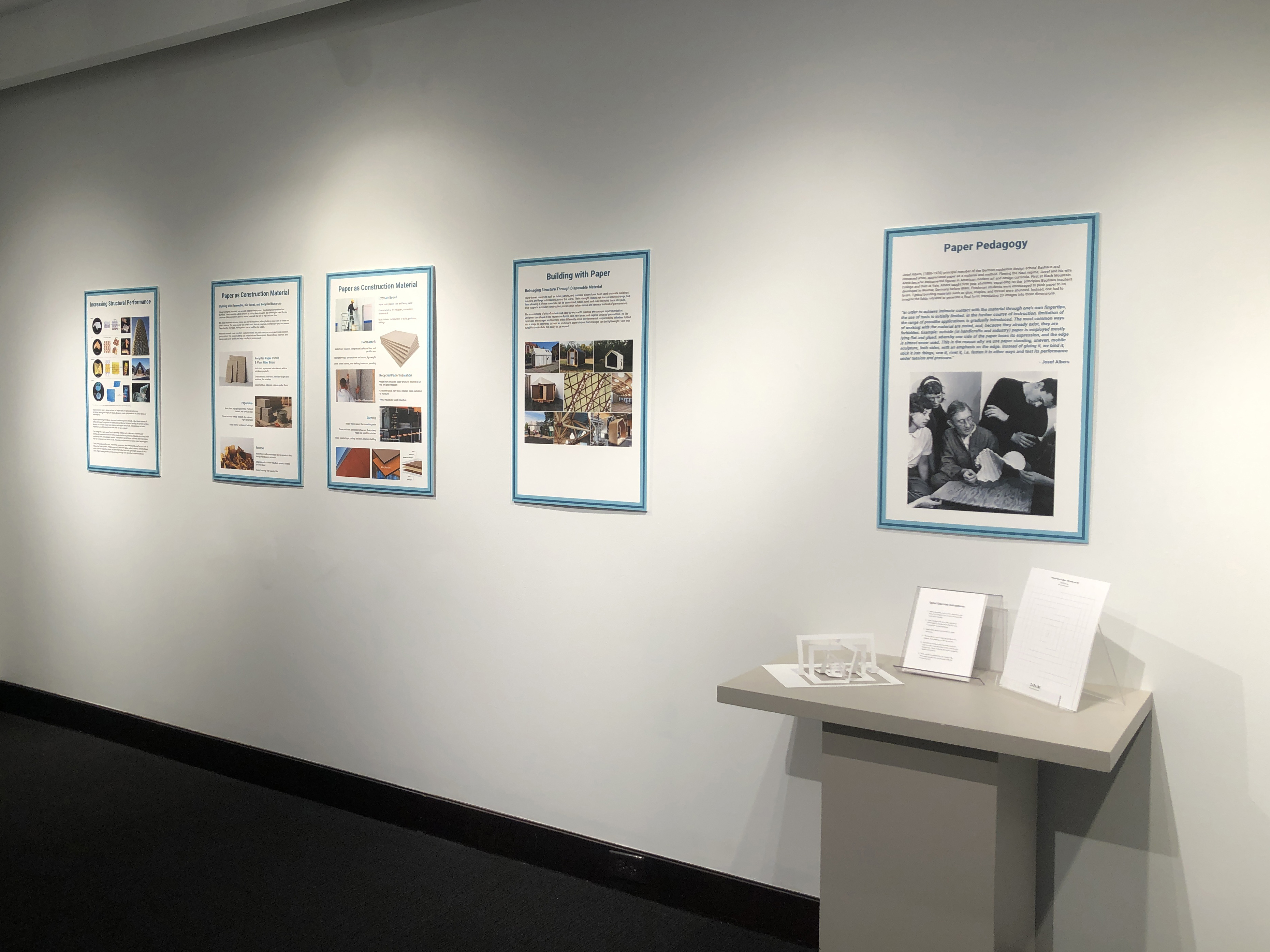 A gallery view of five exhibition boards; to the right is a pedestal holding a DIY paper model. 