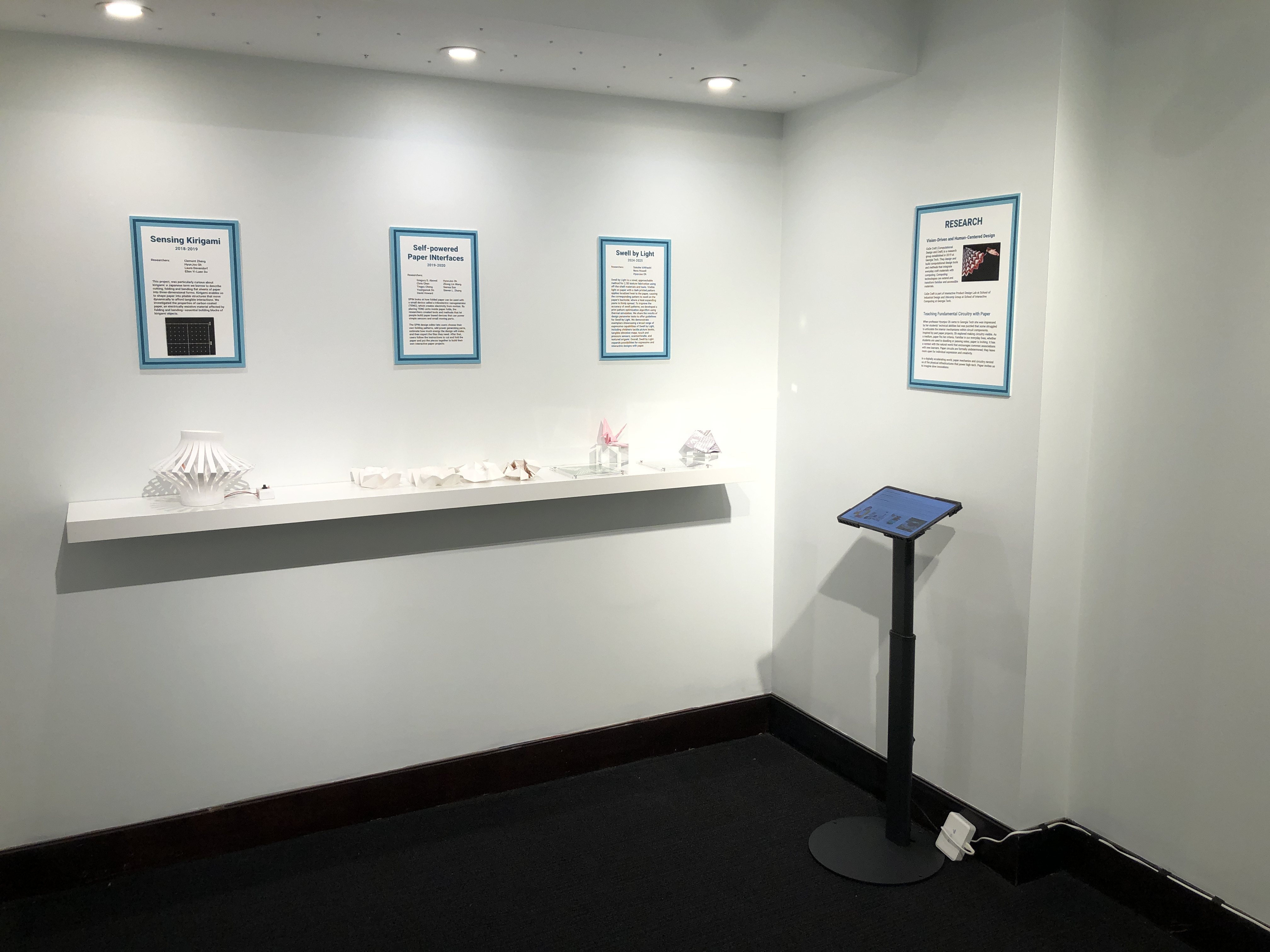 A gallery view of four exhibition boards, mounted above a shelf holding several paper models. 