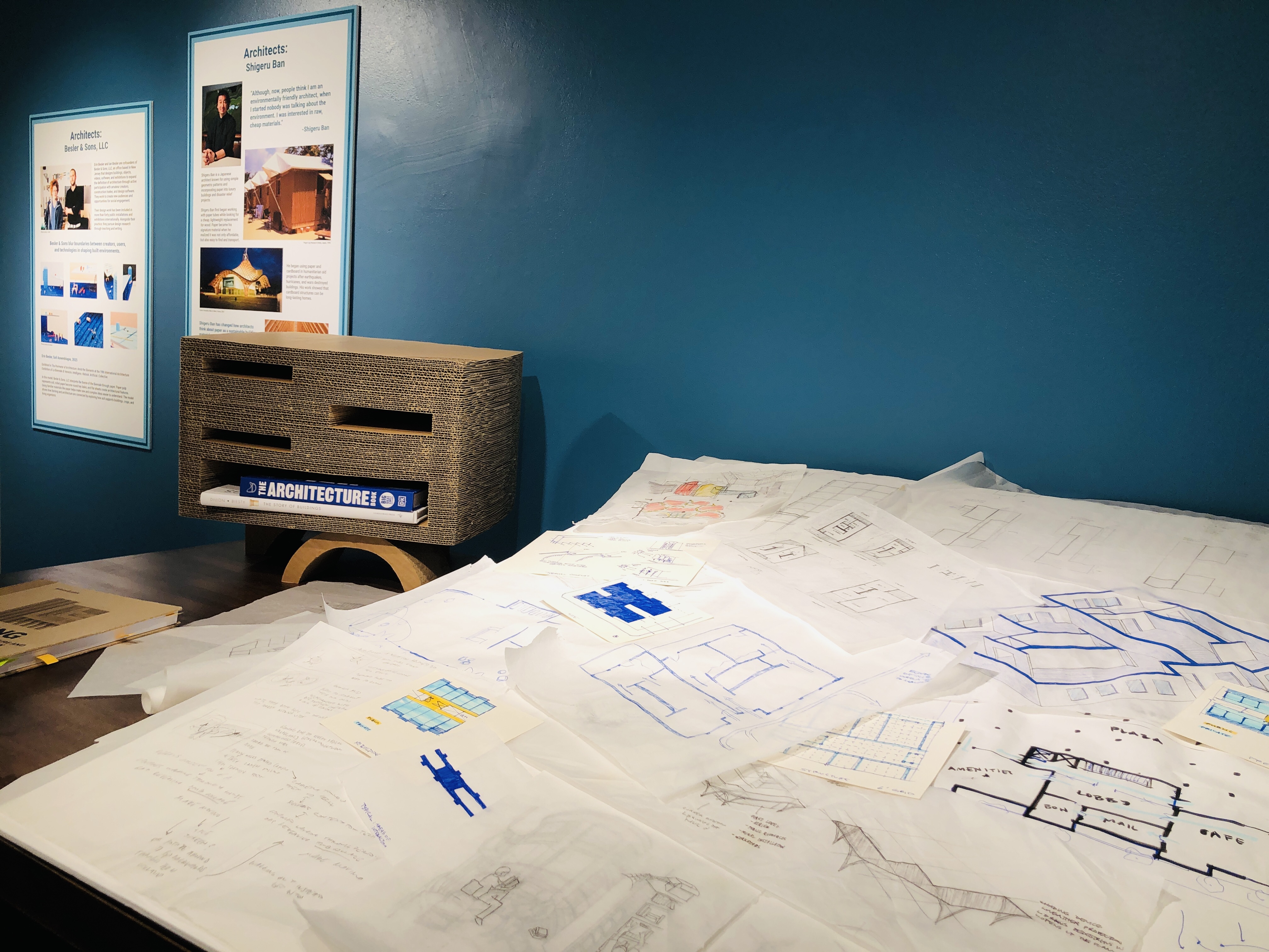 A closeup view of the architect’s table display: several hand drawings on trace paper are displayed, in the background are two exhibition boards. 
