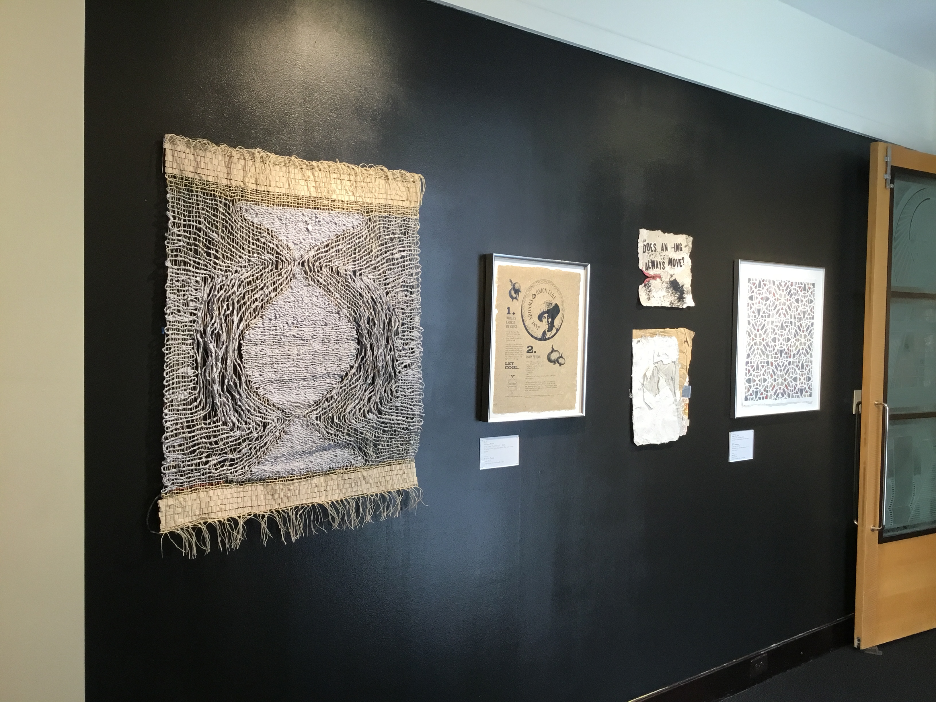 Five artworks on a black wall at the entry into the gallery. First image looks like a woven rug, the second image is a small brown sheet of paper with a portrait printed on it and next to that are abstract pieces made from recycled paper. The last image is a mosaic geometric design.