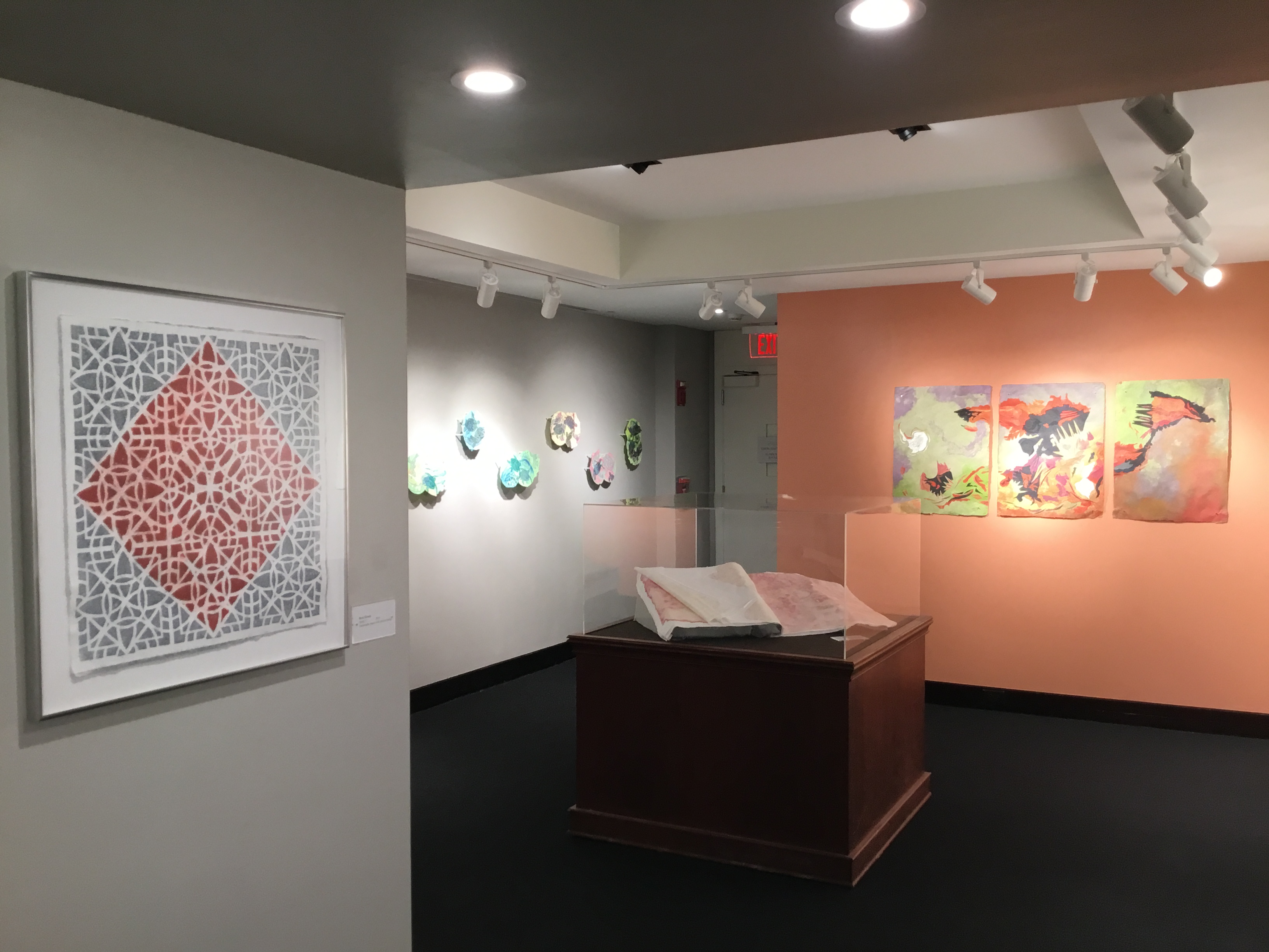 The front and left sides of the back room. On the far left is a piece of paper with a patterned design, colored grey on the outside and a red diamond shape on the inside. Behind this pieces are three-dimensional, multi-color pieces of paper, One the back wall are three marbled pieces of paper.