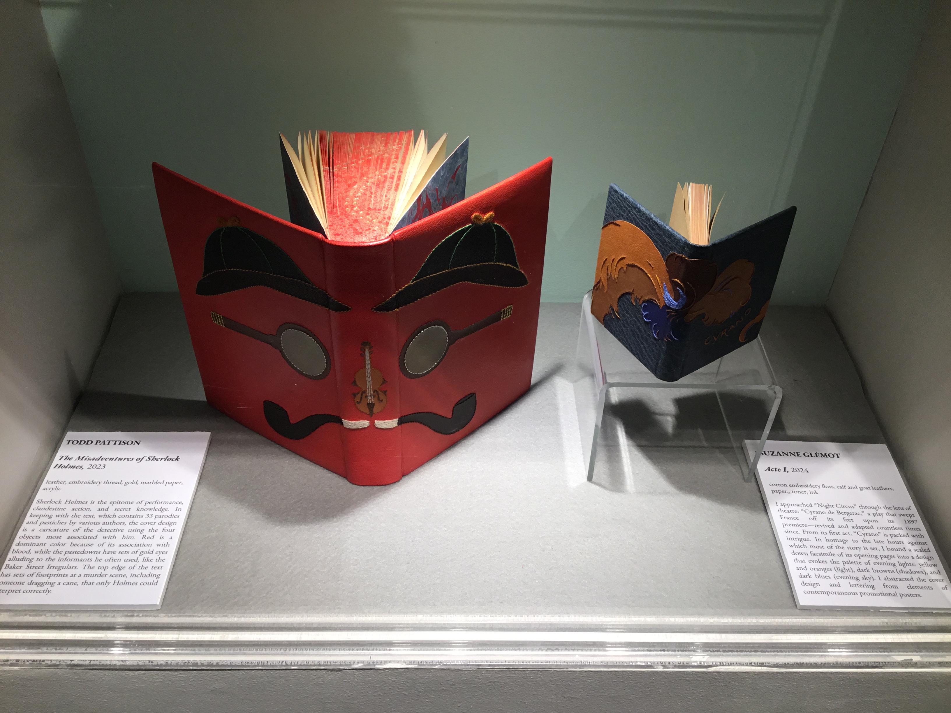 View of a built-in display case in the front gallery with two art books on display. On the left is Todd Pattison’s The Misadventures of Sherlock Holmes, a red leather book with black deerstalker hats, magnifying glasses, smoking pipes and a violin arranged as a face, along the front and back covers. To the right is Suzanne Glemot’s Acts I, a blue leather book with plume-like appliqués in tan, brown, orange and blue, and the word Cyrano embroidered on the front cover.