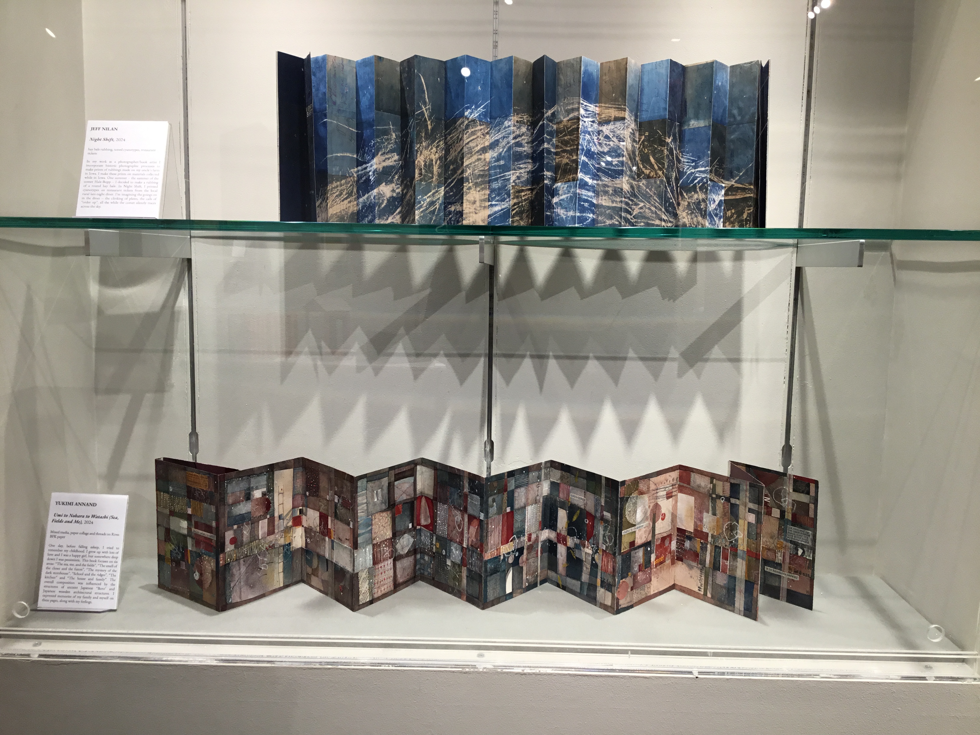 View of a built-in display case in the front gallery. On the top shelf is Jeff Nilan’s Night Shift, a blue accordion book with the imprint of a round hay bale. On the bottom shelf is Yukimi Annand’s Umi to Nohara to Watashi, an open accordion book of a patchworked paper collage in blues, greens, reds, and many shades in between. 