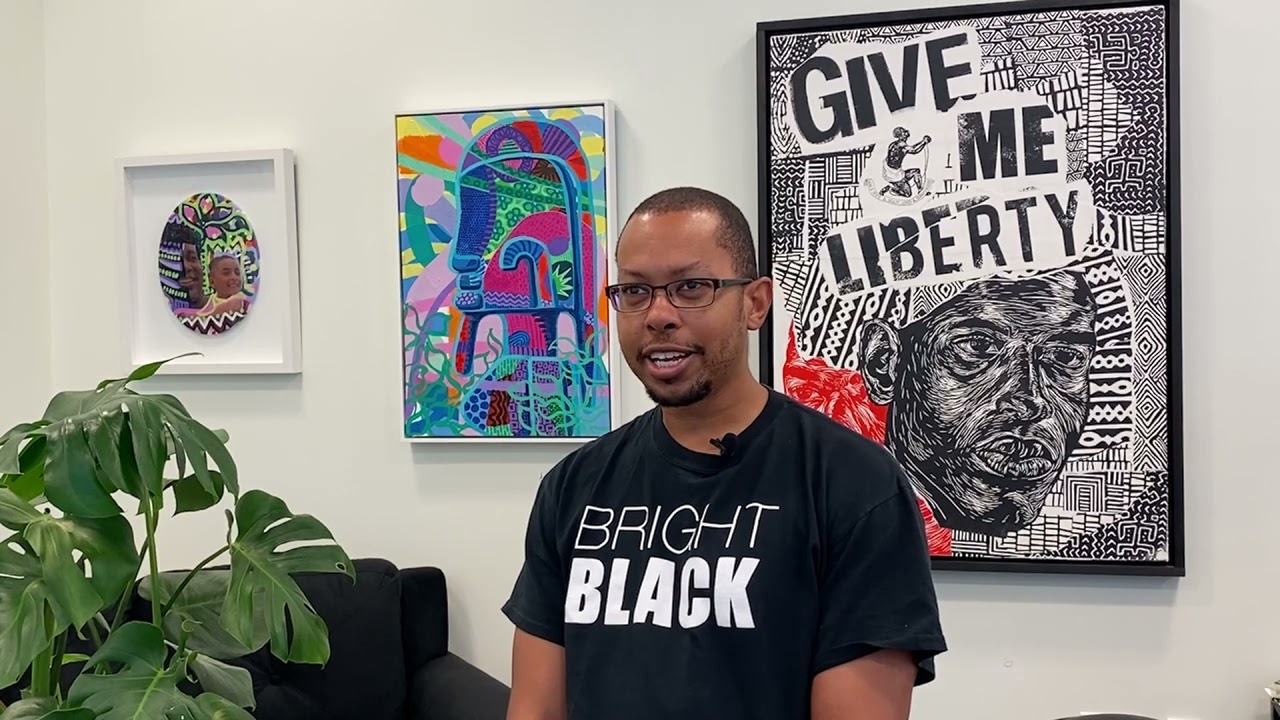 Jamaal Barber, an African-American man with short cropped head and glasses, stands in front of his artwork mounted on the wall