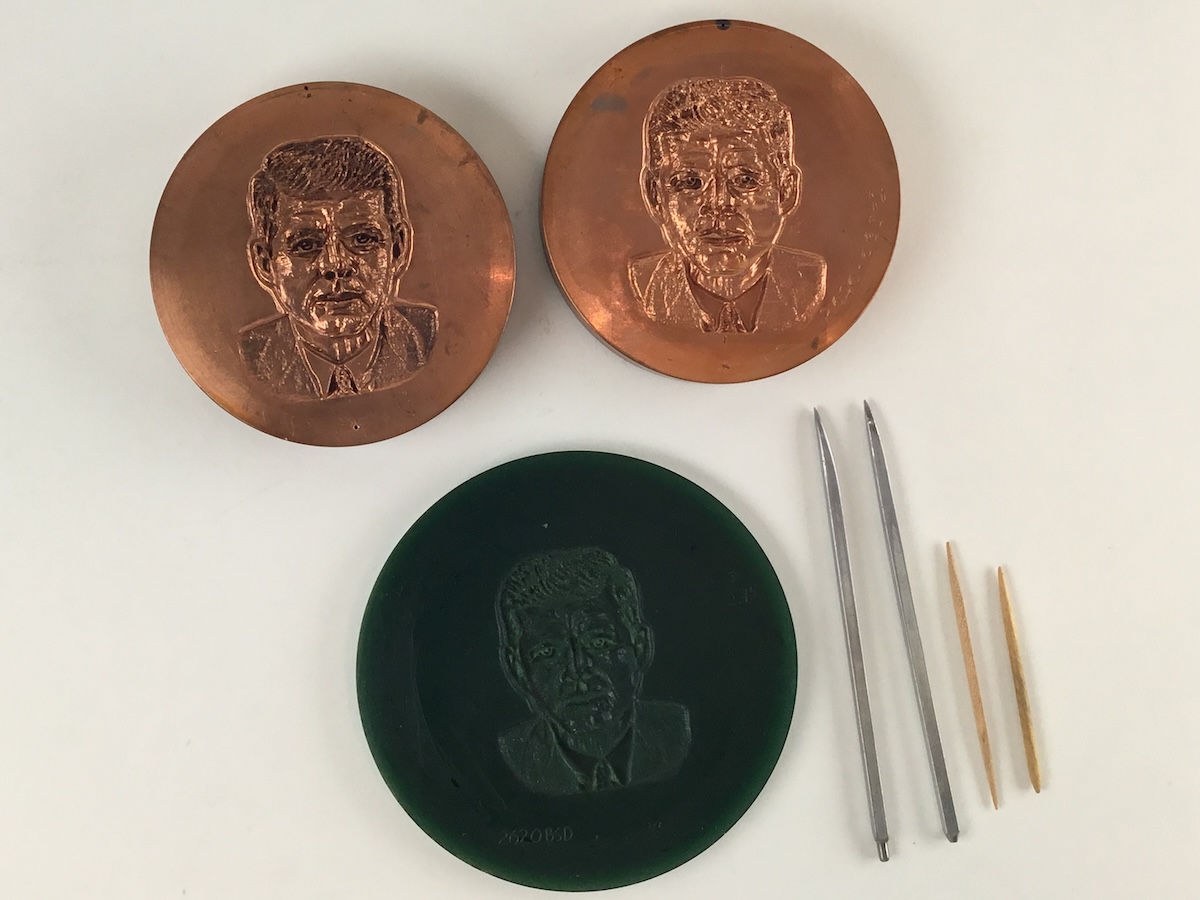 Two copper embossing plates above a dark green silicon mold on a white background. To the right of the plates are the four thin tools used to carve, two silver metal and two wooden sticks.