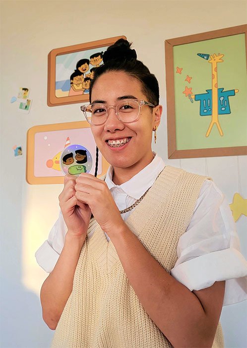 Colored photo of Jalysa Leva in front of a wall of her framed artwork and holding a sticker with characters from her animated series. Jalysa is an Filipino-American woman wearing glasses and a cream sweater vest over a short sleeve white button up shirt. Her hair is shaved on the sides with the long hair on top tied in a top knot.