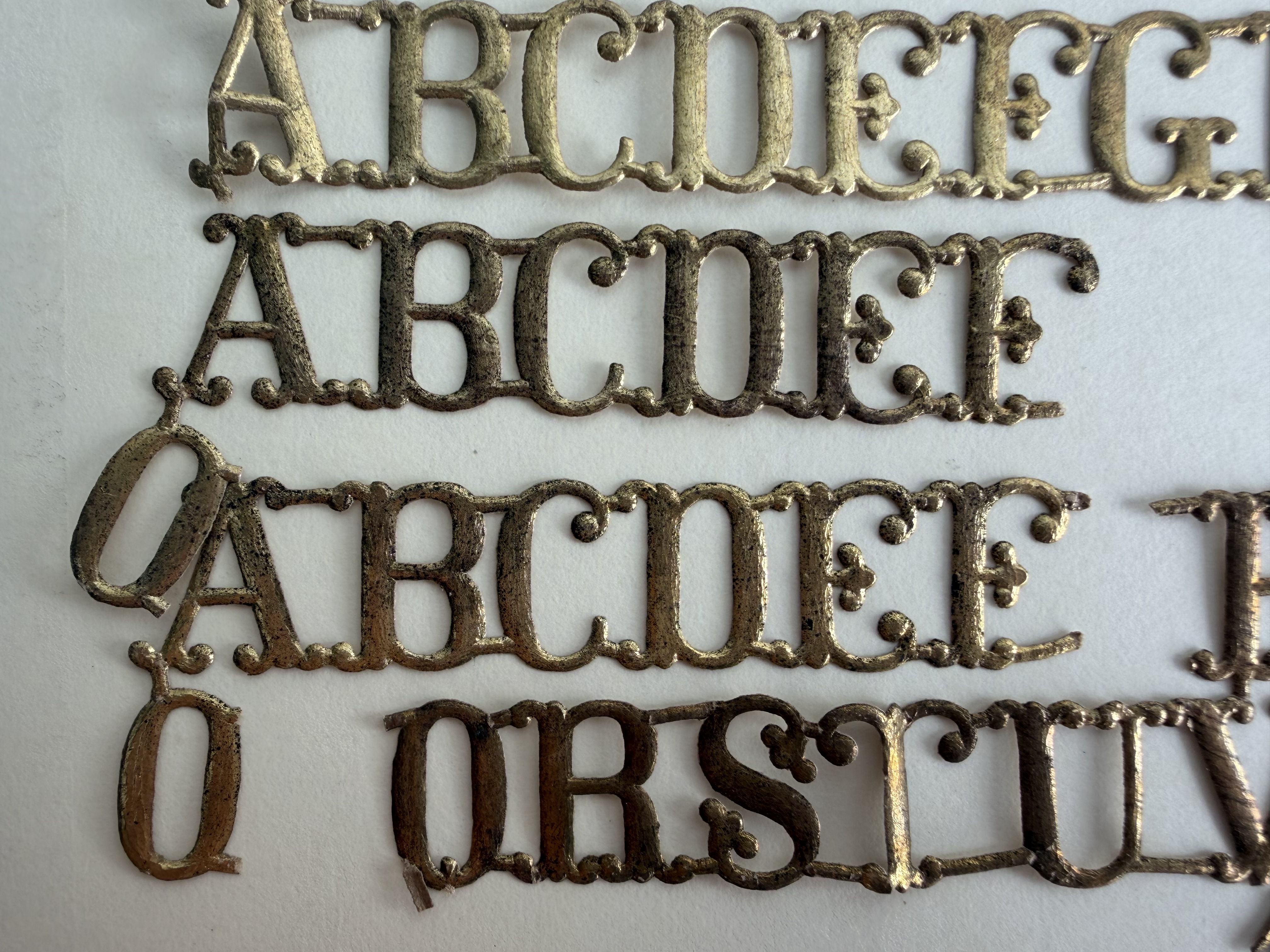 Bronze colored paper shaped like letters A-G and Q-V on white background. They are attached to each other in straight horizontal lines; some lines are shorter than others.