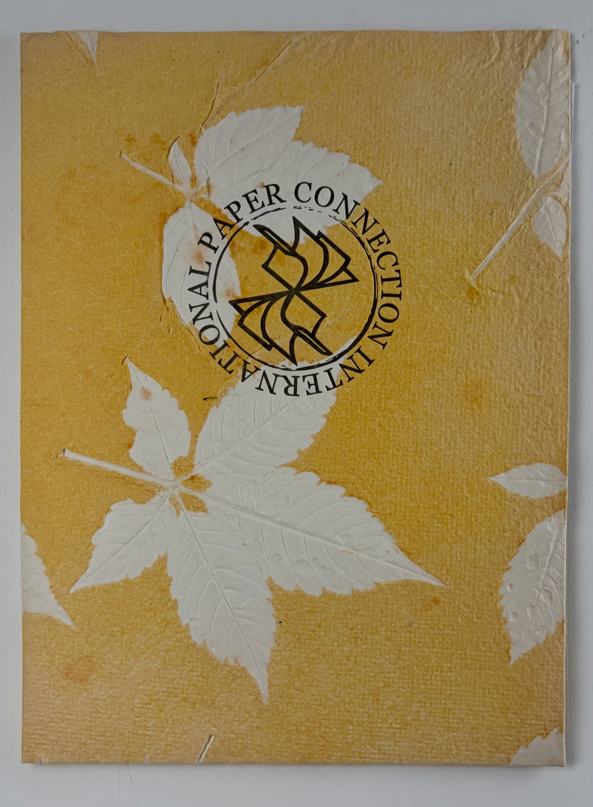 Yellow folder with large white leaf imprints. There is a circle logo in the middle with “Paper Connection International around it in black font.