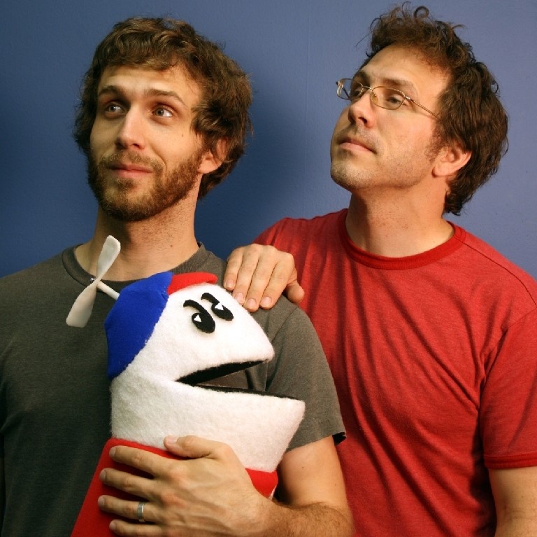 Three-quarter shot of Matt and Mike Chapman standing in front of a blue background looking off toward the upper left corner of the photo. Matt is holding a plush doll of their animated character Homestar Runner. The character is a white figure without a nose, wearing a blue beanie with a propeller on top, a red shirt with a white star and no arms 