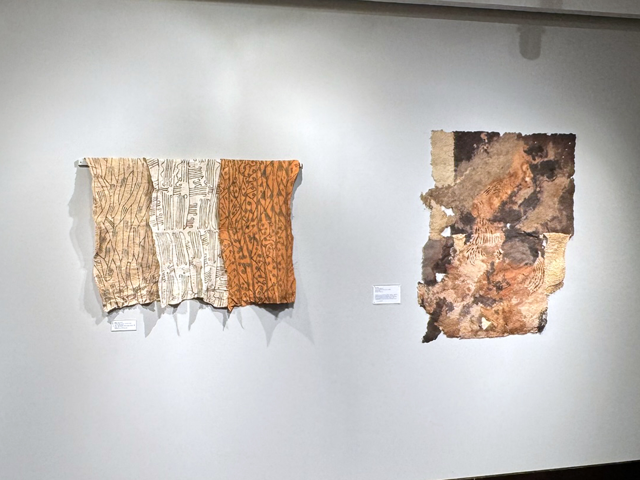 View of two central African barkcloth pieces. On the left, there are three patterned pieces of overlapping barkcloth in tan, white and orange. On the right is an irregularly shaped section of barkcloth with tan, rust, and deep brown splotches.