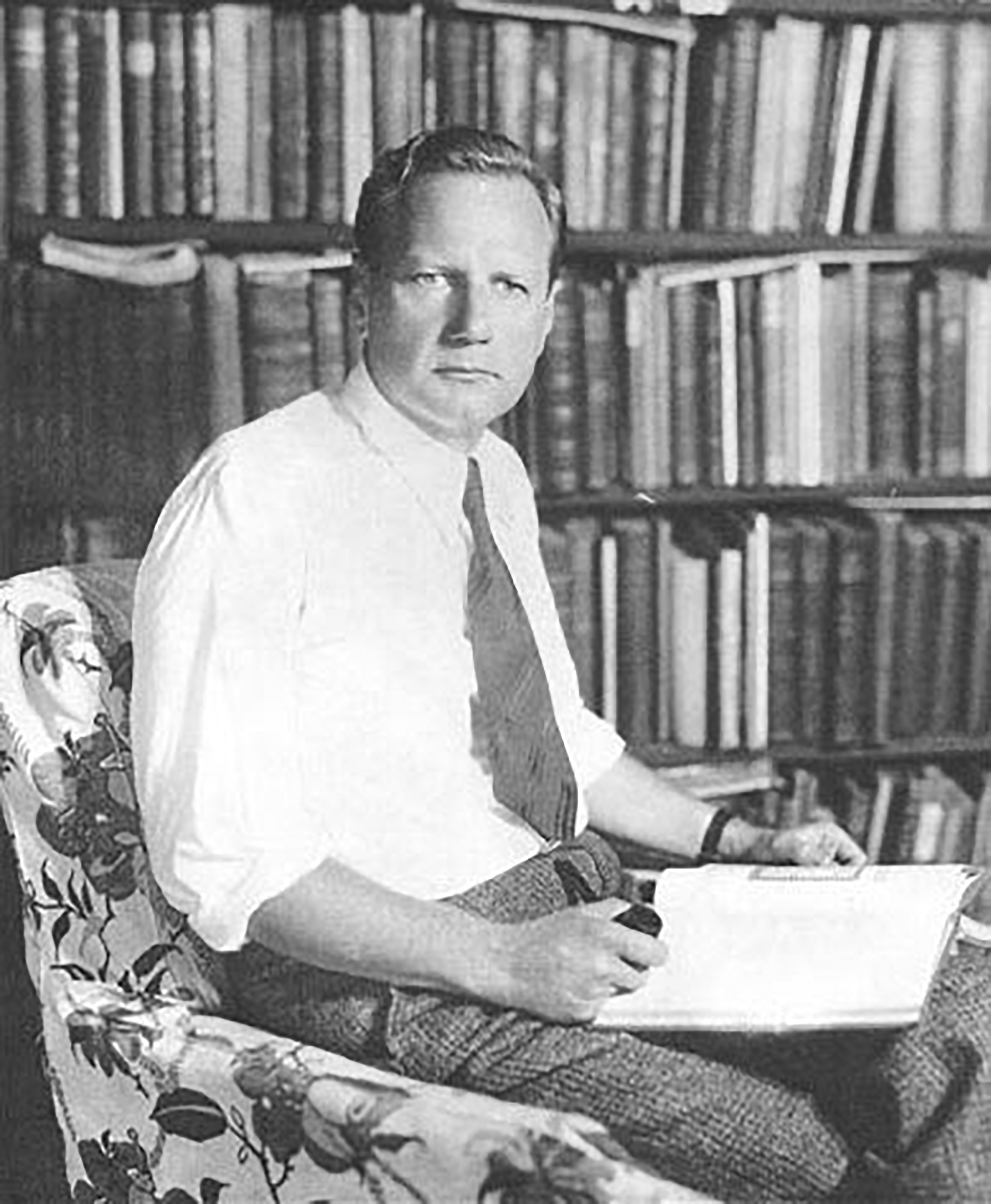 Black and white photo of Dard Hunter sitting in his library. Dard, a caucasian  male, is wearing a tie and a white dress shirt with the sleeves rolled up. He sits in front of a background of bookshelves in a white chair with a floral pattern and is facing the camera with a book on his lap and a pipe in his right hand.