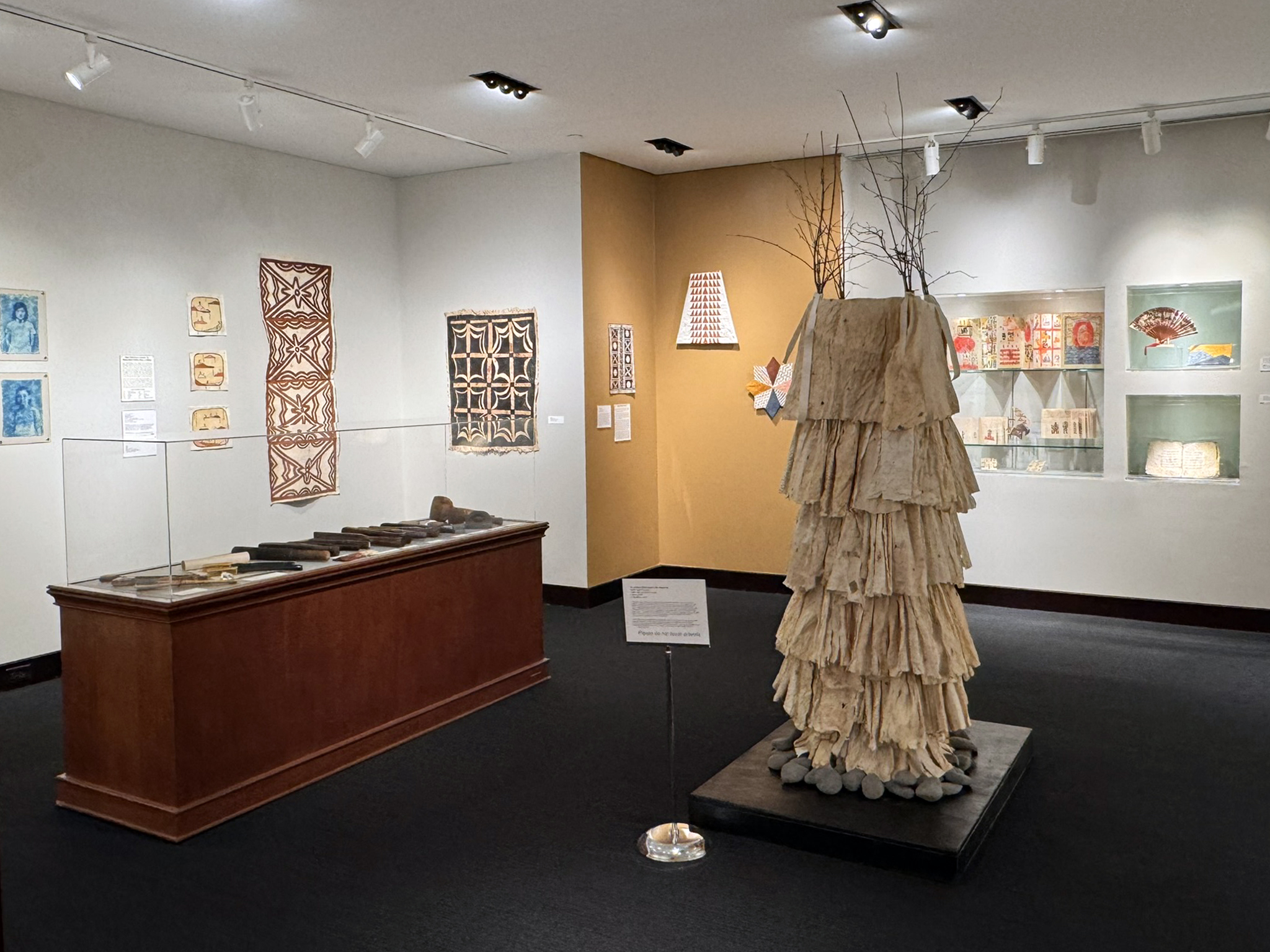 Overview of the front gallery. In the foreground is a large sculpture of layered tiers of light brown bark paper surrounded by a ring of smooth gray stones. To the left is a large display case with several bark beaters. Other various art pieces are on the walls in the background.