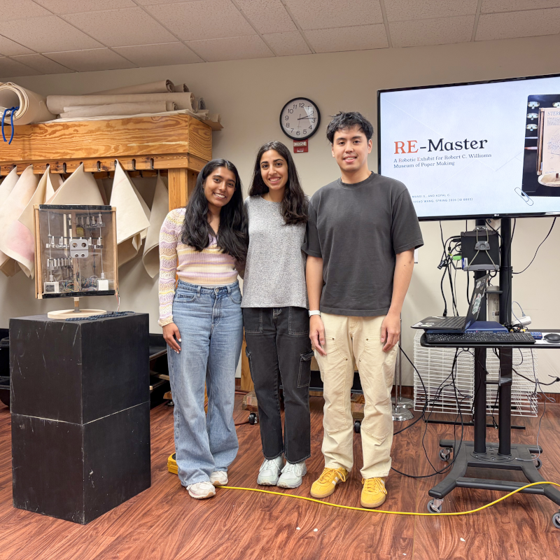 Three students pose beside their robotic exhibit and screen titled RE-Master at a paper museum presentation