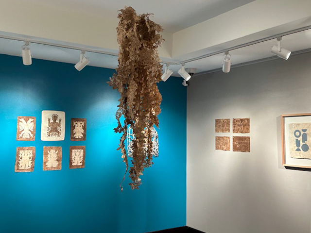 View of the back gallery, in the foreground a sculpture hangs from the ceiling, a mass of wavy strips of leaf-like garland. In the background, the left wall is painted bright blue and displays a series of six Otomi spiritual figure papercuts on bark paper. On the right wall, a set of four animal drawings on bark paper.