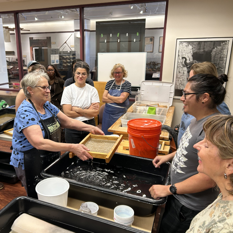 A papermaking instructor lifts a mould from a vat of pulp while a group watches closely in a workshop studio.