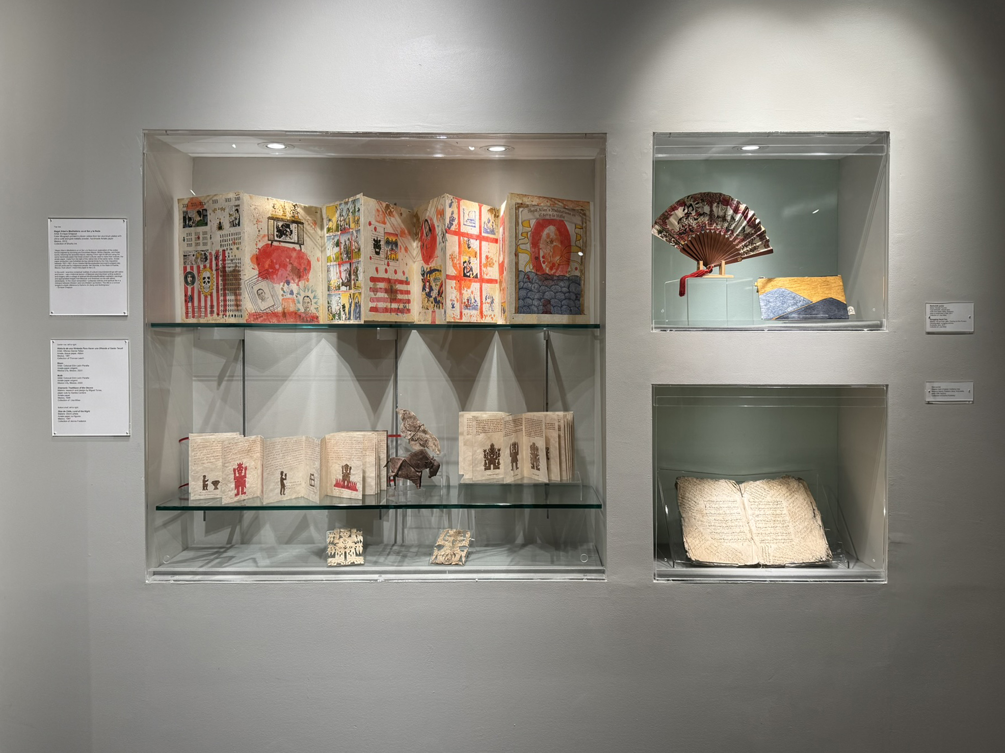 View of a built-in display case in the front gallery. On the left is a collection of accordion books, origami and papercut figures, all on amate paper. On the top right is an unfolded red and white paper fan, next to a barkcloth purse depicting blue mountains over a yellow background. On the bottom right is an open manuscript made of light brown bark paper.