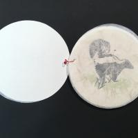 A thin, circular sheet of translucent paper with floral and plant-like embeds covers a painted image of a skunk in a patch of grass.