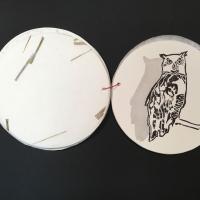 A circular sheet of paper with a black and white painted image of a Great Horned Owl standing on a branch.