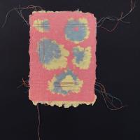 A red piece of handmade paper with deckled edges and blue and yellow patches, as well as red, blue, and yellow thread running horizontally and vertically around the border.
