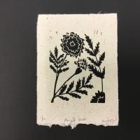 This piece, entitled “Marigold Harvest”, is a rectangular sheet of white handmade paper with small black specks and raw edges. In the center of the paper is a black ink-print of a marigold.