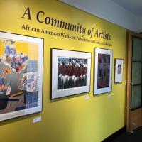 An image of the title wall of the gallery, with large, deep purple text on a yellow background with four framed pieces underneath. The text reads “A Community of Artists: African American Works on Paper from the Cochran Collection.”