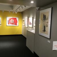 An image of the right side of the corridor and the yellow back wall of the second gallery, with framed pieces hanging all along the walls. 
