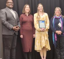 4 people standing facing outward and smiling. One is holding a plaque. 